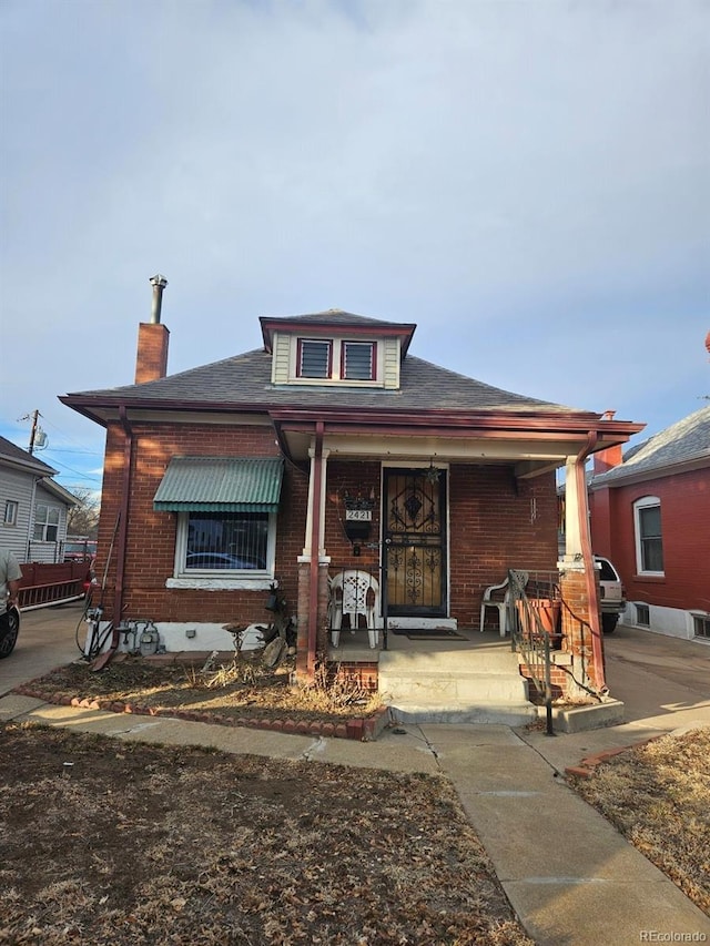 bungalow with a porch, brick siding, a chimney, and a shingled roof