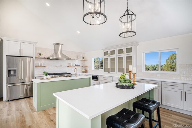 kitchen featuring open shelves, light countertops, appliances with stainless steel finishes, a kitchen island with sink, and wall chimney range hood