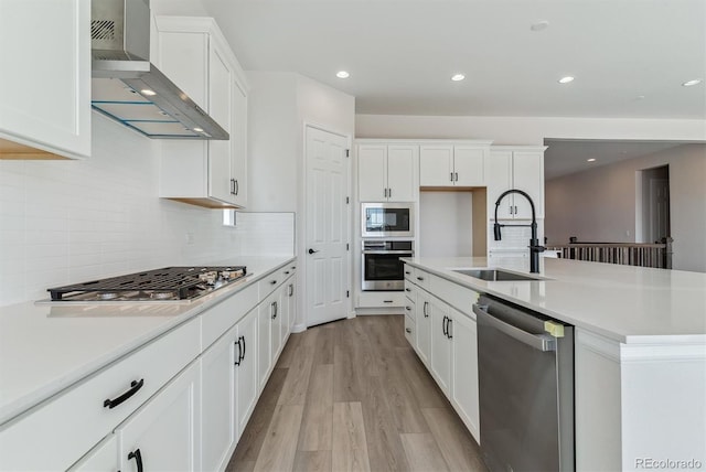 kitchen with stainless steel appliances, a sink, white cabinets, wall chimney exhaust hood, and light wood finished floors