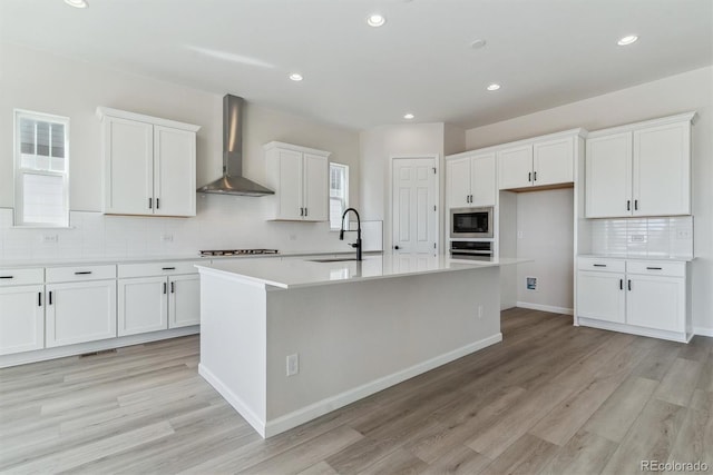 kitchen with white cabinets, wall chimney exhaust hood, a kitchen island with sink, stainless steel appliances, and a sink