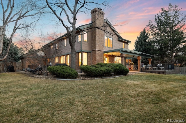 back of property at dusk featuring a yard, brick siding, a chimney, and fence