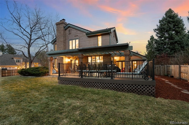 back of property at dusk with a wooden deck, a lawn, a fenced backyard, and a chimney