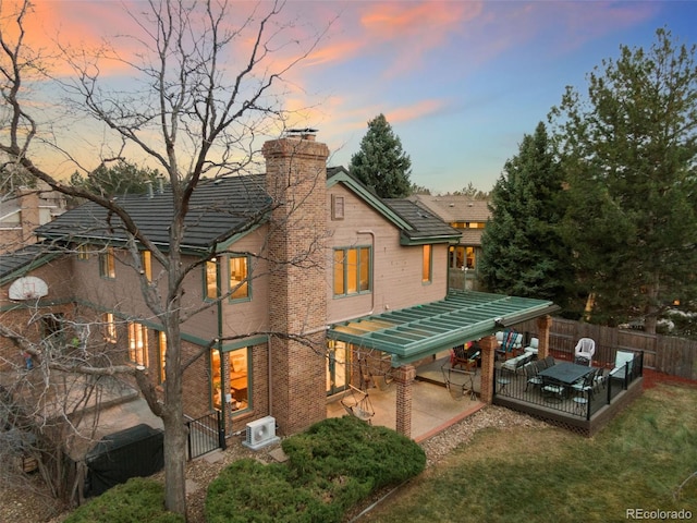 back of house at dusk featuring a lawn, a patio, fence, brick siding, and a chimney