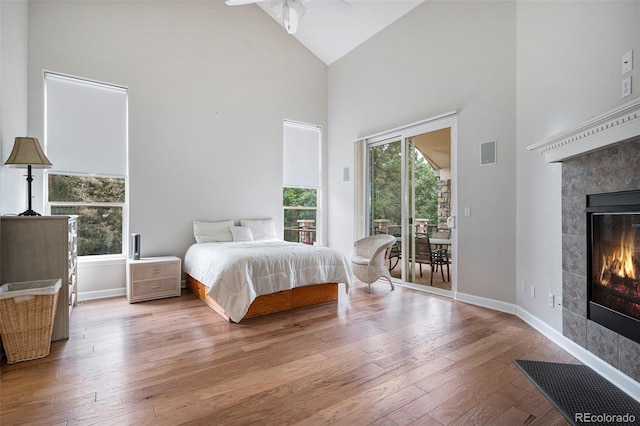 bedroom with high vaulted ceiling, access to outside, a tiled fireplace, hardwood / wood-style floors, and baseboards