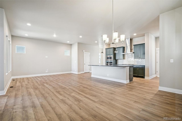 kitchen with stainless steel microwave, wall chimney exhaust hood, light hardwood / wood-style flooring, an island with sink, and decorative light fixtures