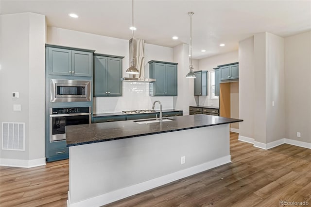 kitchen with sink, wall chimney exhaust hood, an island with sink, pendant lighting, and appliances with stainless steel finishes