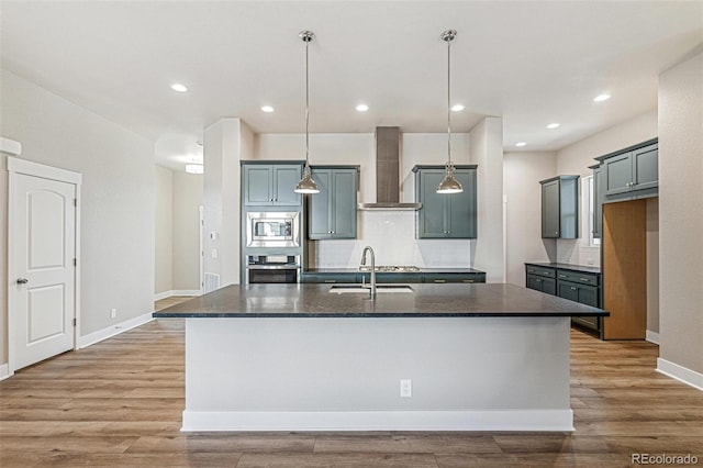 kitchen featuring decorative light fixtures, stainless steel appliances, wall chimney exhaust hood, and an island with sink