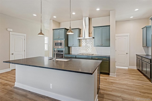 kitchen featuring pendant lighting, a kitchen island with sink, wall chimney range hood, sink, and appliances with stainless steel finishes