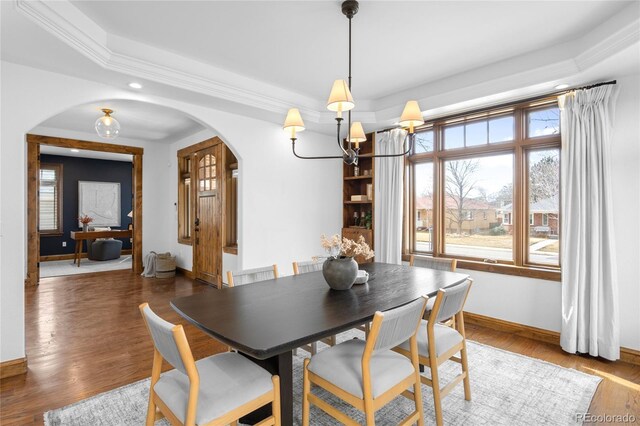 dining area with arched walkways, wood finished floors, baseboards, a raised ceiling, and an inviting chandelier