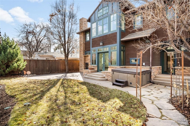 back of property with a hot tub, a patio, stone siding, a chimney, and fence