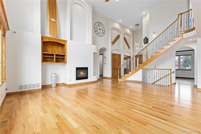 unfurnished living room with light wood finished floors, visible vents, stairway, a high ceiling, and a glass covered fireplace