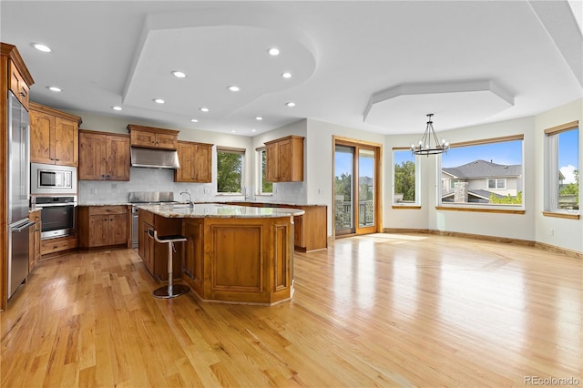 kitchen with a breakfast bar area, brown cabinetry, light stone countertops, under cabinet range hood, and built in appliances