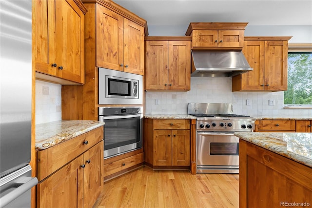 kitchen with light stone counters, brown cabinets, stainless steel appliances, backsplash, and wall chimney exhaust hood