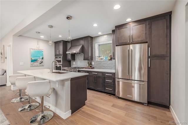 kitchen featuring wall chimney range hood, tasteful backsplash, decorative light fixtures, a kitchen island with sink, and appliances with stainless steel finishes