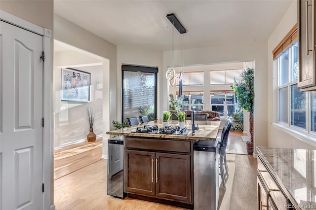 kitchen with stainless steel gas stovetop, dark brown cabinetry, hanging light fixtures, light wood-type flooring, and an island with sink