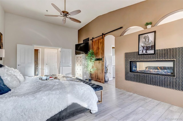 bedroom featuring ceiling fan, a barn door, lofted ceiling, a fireplace, and light wood-type flooring