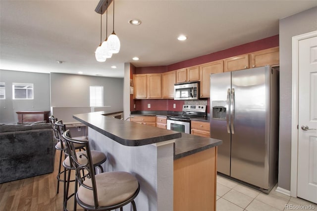kitchen with light brown cabinets, dark countertops, open floor plan, a kitchen bar, and appliances with stainless steel finishes