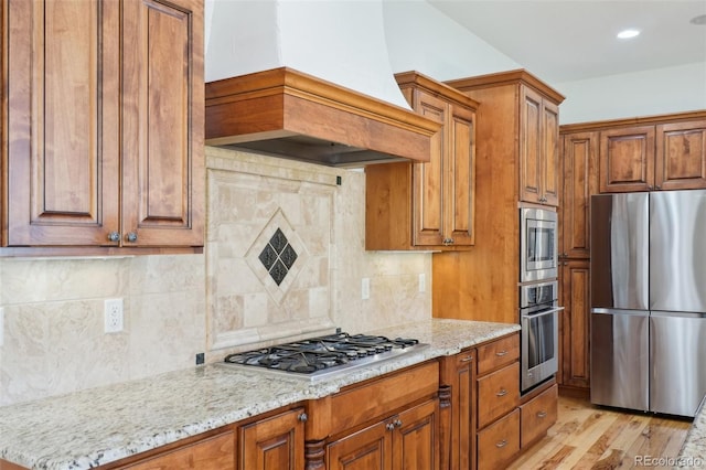 kitchen with appliances with stainless steel finishes, custom exhaust hood, brown cabinetry, and light stone countertops