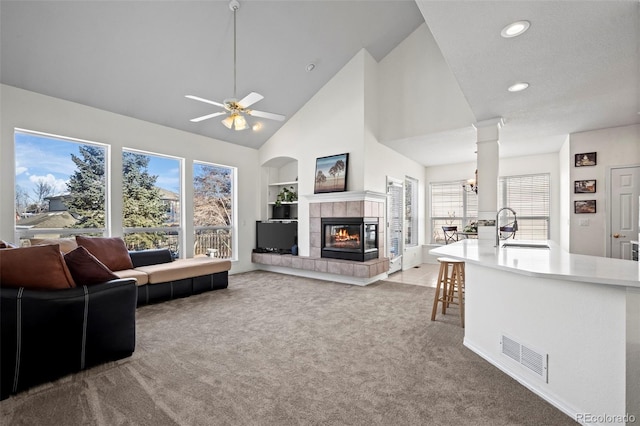 carpeted living room with high vaulted ceiling, ceiling fan with notable chandelier, sink, built in features, and a tiled fireplace