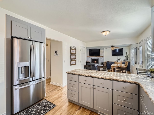 kitchen featuring gray cabinets, a fireplace, stainless steel fridge, light hardwood / wood-style floors, and light stone counters