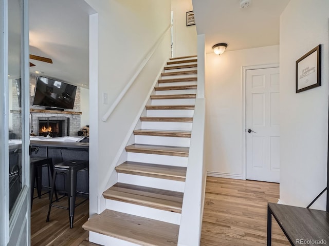 stairway with ceiling fan, wood-type flooring, and a brick fireplace