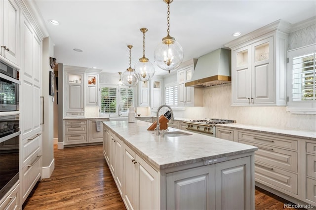 kitchen featuring decorative light fixtures, premium range hood, light stone countertops, an island with sink, and glass insert cabinets