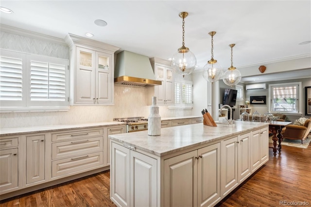 kitchen with a wall unit AC, an island with sink, glass insert cabinets, hanging light fixtures, and wall chimney range hood