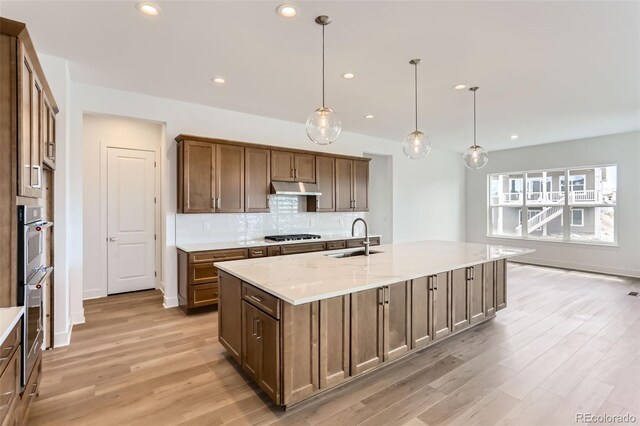 kitchen with a sink, under cabinet range hood, light wood-type flooring, decorative backsplash, and an island with sink