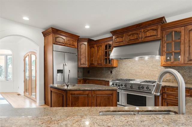 kitchen featuring ventilation hood, stone counters, arched walkways, decorative backsplash, and appliances with stainless steel finishes