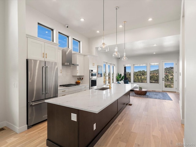 kitchen featuring pendant lighting, wall chimney range hood, appliances with stainless steel finishes, a kitchen island with sink, and white cabinets