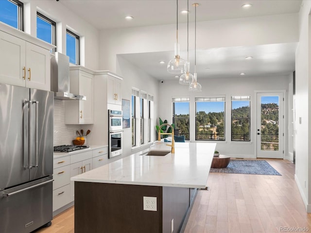 kitchen featuring sink, appliances with stainless steel finishes, white cabinets, a center island with sink, and wall chimney exhaust hood