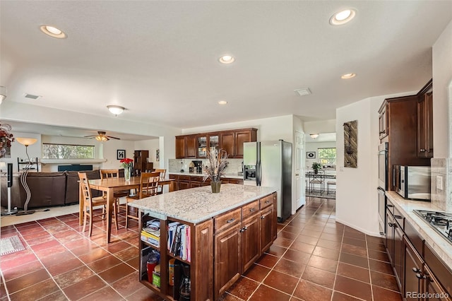 kitchen with a kitchen island, appliances with stainless steel finishes, dark tile patterned flooring, ceiling fan, and light stone counters