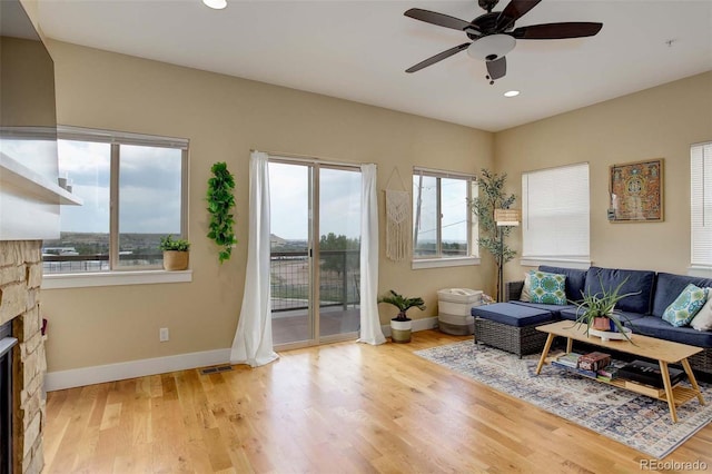 living room featuring light wood-type flooring, a stone fireplace, ceiling fan, and a wealth of natural light