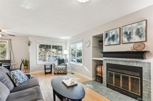 living room with a tile fireplace, ceiling fan, built in features, a textured ceiling, and wood-type flooring