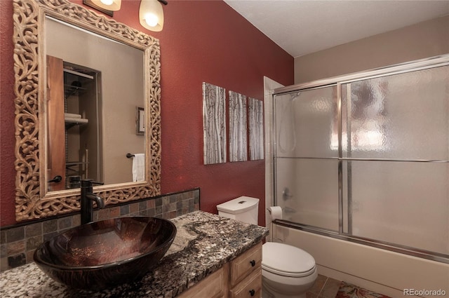 full bathroom featuring shower / bath combination with glass door, vanity, a textured wall, backsplash, and toilet