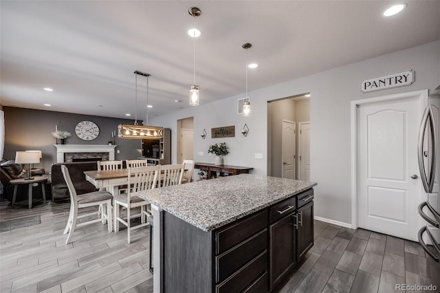 kitchen featuring light stone counters, stainless steel refrigerator, hanging light fixtures, dark brown cabinetry, and a kitchen island