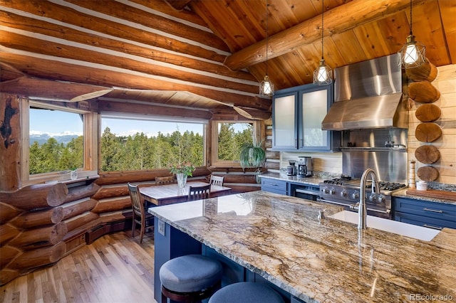 kitchen featuring a breakfast bar, hanging light fixtures, vaulted ceiling with beams, light stone counters, and wall chimney exhaust hood