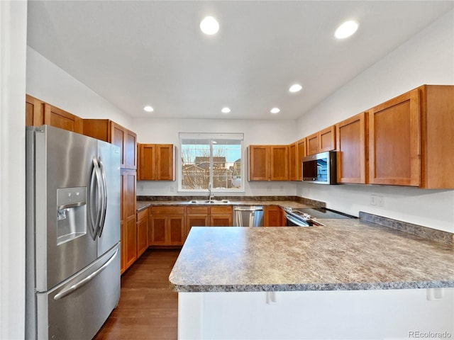 kitchen with dark wood-style flooring, appliances with stainless steel finishes, brown cabinetry, a sink, and a peninsula