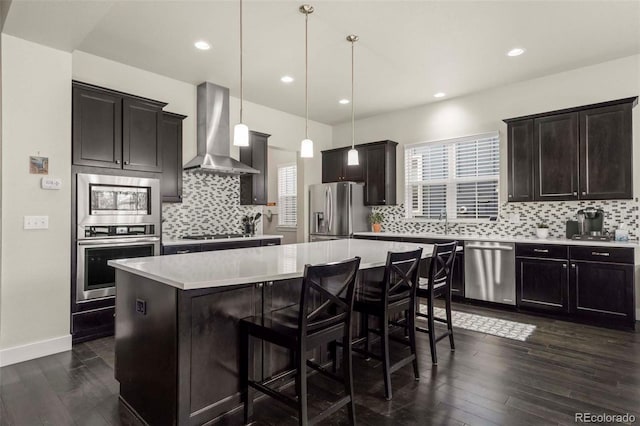 kitchen featuring stainless steel appliances, dark wood-type flooring, wall chimney range hood, a kitchen island, and hanging light fixtures