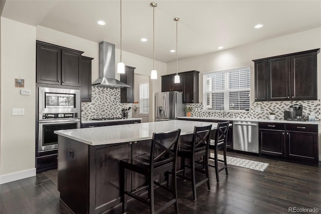 kitchen with wall chimney range hood, appliances with stainless steel finishes, dark wood finished floors, a center island, and backsplash