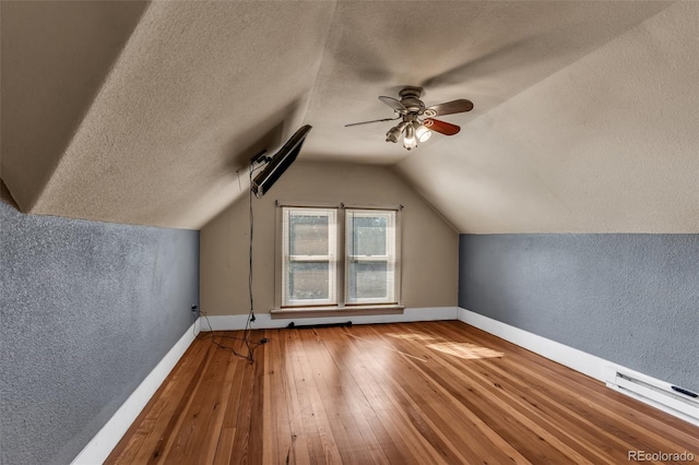 additional living space featuring vaulted ceiling, a textured ceiling, wood-type flooring, ceiling fan, and a baseboard radiator