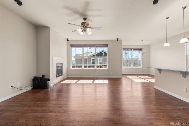 unfurnished living room with ceiling fan with notable chandelier, wood finished floors, a glass covered fireplace, and baseboards