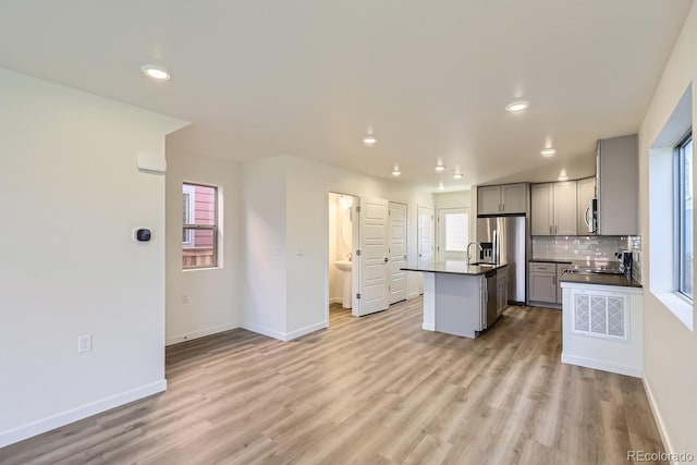 kitchen featuring gray cabinets, backsplash, a kitchen island with sink, stainless steel appliances, and light wood-type flooring