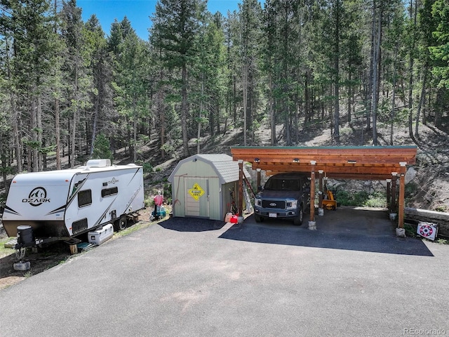 8340 London Ln, Conifer, CO 80433 - view of parking / parking lot featuring a storage unit, a detached carport, asphalt driveway, and a wooded view