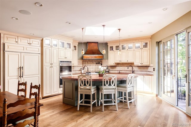 kitchen with hanging light fixtures, premium range hood, cream cabinets, and a kitchen island with sink