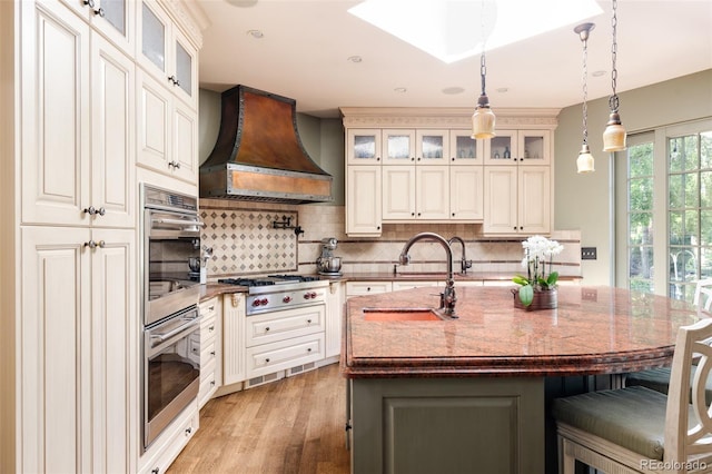 kitchen with hanging light fixtures, a center island with sink, appliances with stainless steel finishes, custom range hood, and light stone countertops