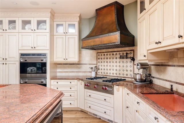 kitchen with sink, custom exhaust hood, light stone counters, stainless steel appliances, and backsplash