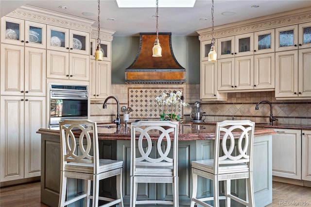 kitchen featuring premium range hood, stainless steel oven, a center island with sink, and cream cabinets