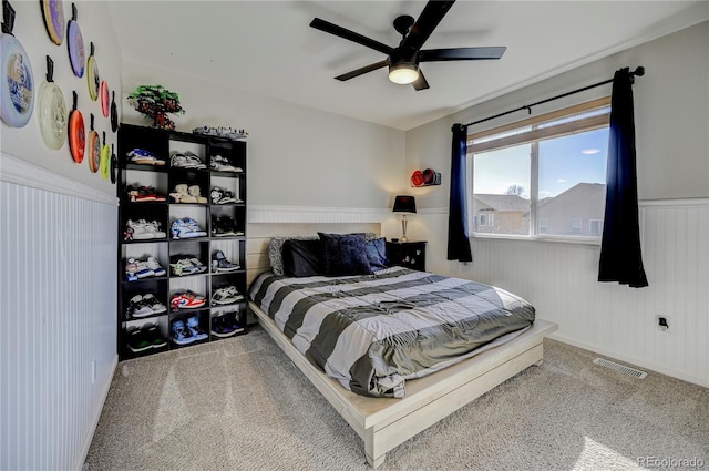 carpeted bedroom with visible vents, a wainscoted wall, and ceiling fan