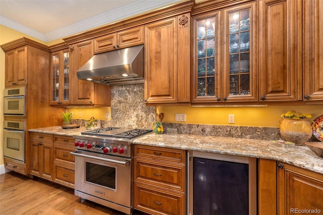 kitchen with wine cooler, glass insert cabinets, appliances with stainless steel finishes, brown cabinets, and exhaust hood